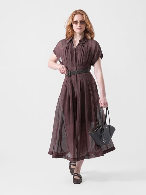 Una donna con un vestito marrone trasparente con cintura, occhiali da sole e sandali, che porta una borsa nera, cammina con sicurezza davanti a uno sfondo bianco semplice.