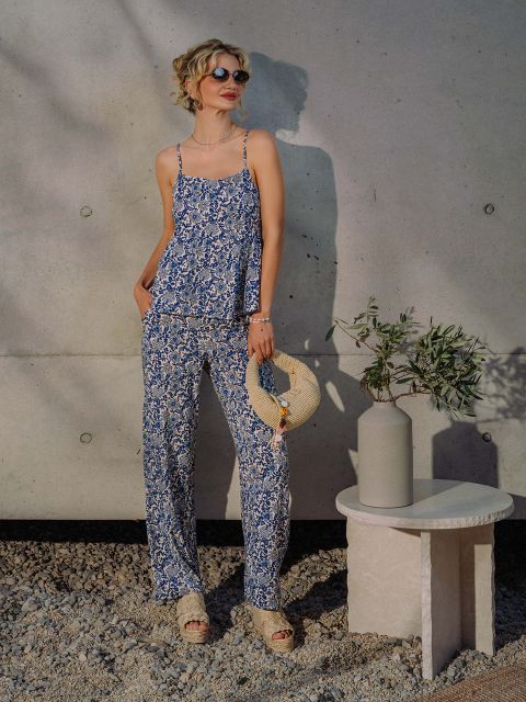 Una donna con occhiali da sole e un vestito blu floreale è in piedi accanto a un muro di cemento, tenendo un cappello di paglia accanto a un vaso con rami d'ulivo su un tavolino.