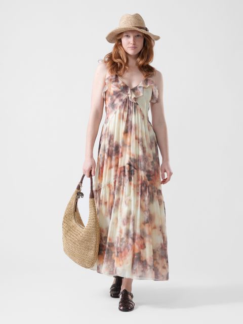 Una donna in un maxi abito fluido e colorato, cappello di paglia e sandali, che tiene in mano una grande borsa intrecciata, si trova davanti a uno sfondo bianco semplice.