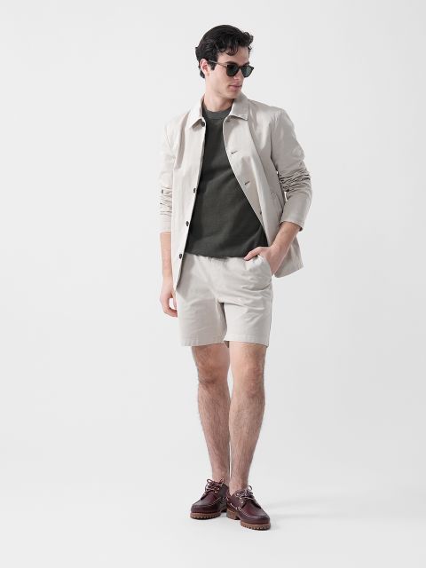 Un uomo con occhiali da sole, giacca beige, camicia verde oliva, pantaloncini beige e scarpe marroni si trova di fronte a un semplice sfondo bianco.