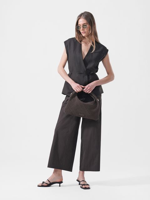 Una donna indossa occhiali da sole, un top nero senza maniche a portafoglio, pantaloni neri a gamba larga e sandali con tacco, e tiene una borsa marrone testurizzata su uno sfondo bianco.