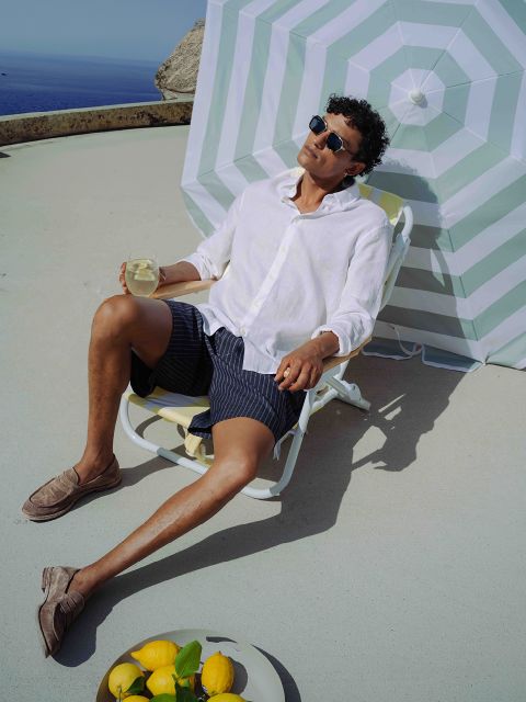 Uomo con occhiali da sole e camicia bianca si rilassa su una sedia a sdraio con una bevanda all'ombra di un ombrellone a righe; limoni e un bicchiere sono a terra nelle vicinanze.