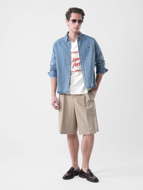 Uomo che indossa occhiali da sole, una camicia di jeans sopra una maglietta grafica, pantaloncini beige al ginocchio e scarpe basse marroni, in piedi davanti a uno sfondo semplice.