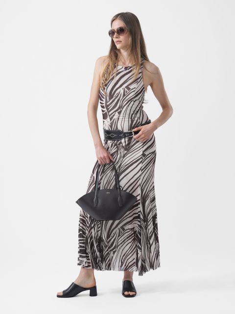 Donna con occhiali da sole, che indossa un maxi abito a fantasia bianco e nero, una cintura nera e sandali neri, e tiene una borsa nera davanti a uno sfondo bianco.