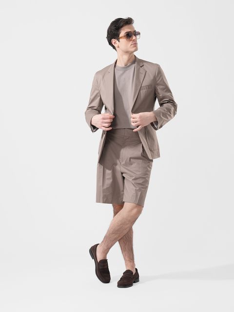 Un uomo che indossa occhiali da sole, un blazer color talpa, pantaloncini coordinati, una maglietta grigia e mocassini marroni, in piedi davanti a uno sfondo bianco semplice.
