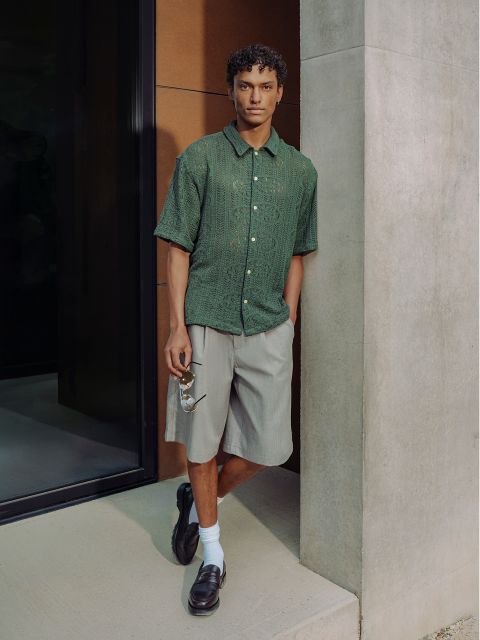 Un uomo con una camicia verde a maniche corte, pantaloncini grigi, calzini bianchi e scarpe nere basse è appoggiato a un muro e tiene in mano un paio di occhiali da sole.
