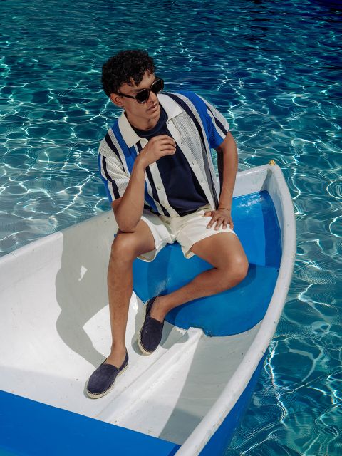 Un uomo con occhiali da sole, camicia a righe e pantaloncini è seduto rilassato in una piccola barca bianco-blu sull'acqua limpida, scintillante e blu.