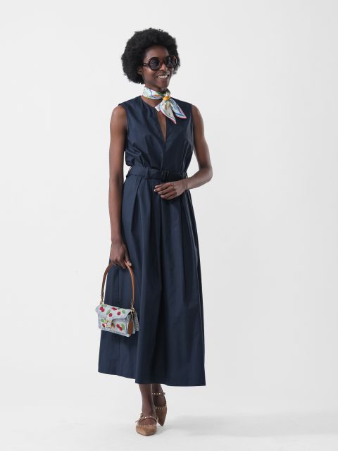 Donna in un vestito blu navy senza maniche, una sciarpa colorata, occhiali da sole, una borsa a fantasia e scarpe marroni, su sfondo bianco.