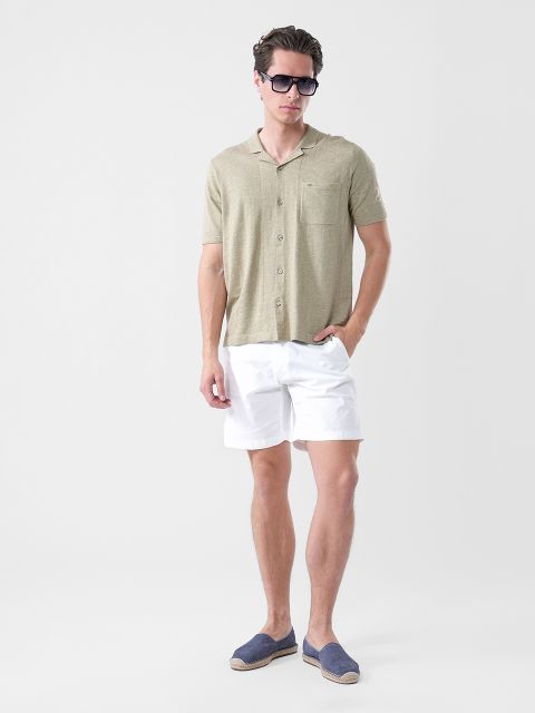 Un uomo che indossa occhiali da sole, una camicia a maniche corte color oliva chiaro, pantaloncini bianchi ed espadrillas blu navy, in piedi davanti a uno sfondo bianco semplice.