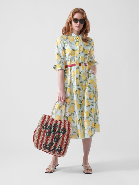 Una donna con un vestito a fantasia di limoni, occhiali da sole e sandali, tiene una borsa a righe rosse e bianche con una scritta, davanti a uno sfondo bianco semplice.