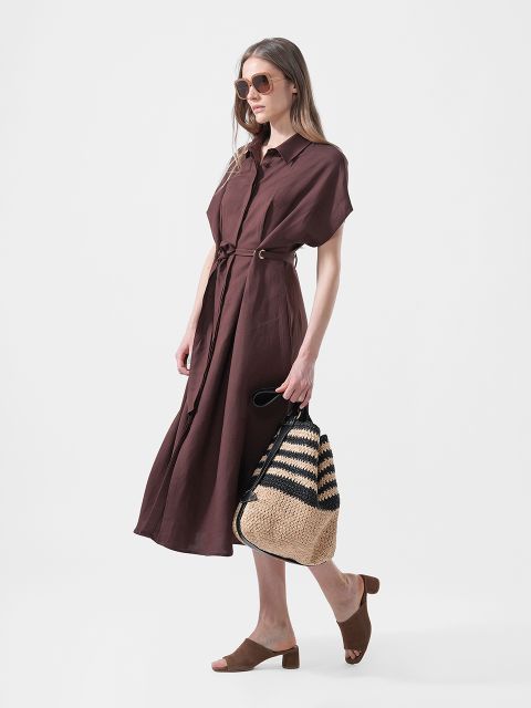 Una donna indossa un vestito midi marrone con cintura, sandali piatti marroni, occhiali da sole e una borsa tote in paglia intrecciata con strisce nere.