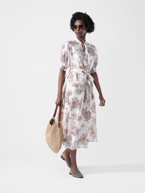 Donna in un vestito midi floreale con fiocco in vita, con occhiali da sole, una borsa intrecciata in mano e mocassini marroni.