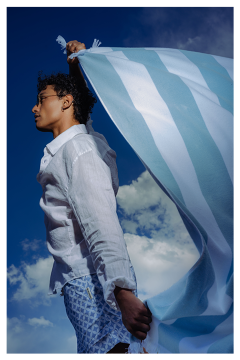 Persona in una camicia bianca, che tiene un tessuto a strisce blu e bianche, davanti a un cielo blu con nuvole.