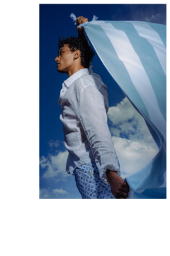 Una persona in camicia bianca e pantaloncini fantasia tiene un panno a strisce blu e bianche davanti a un cielo blu con nuvole.