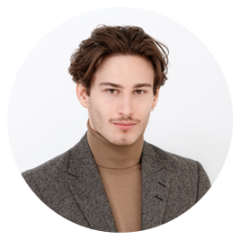 Giovane uomo con capelli corti castani, che indossa un blazer grigio e un dolcevita marrone chiaro, e posa davanti a uno sfondo semplice e chiaro.