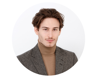 Giovane uomo con capelli corti castani, che indossa un blazer grigio e un dolcevita marrone chiaro, e posa davanti a uno sfondo semplice e chiaro.