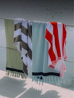 Quattro asciugamani con fantasie e colori diversi sono appesi a una recinzione di corda in riva all'acqua e proiettano ombre sul terreno sottostante.