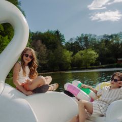 Due bambini con occhiali da sole si rilassano su un pedalò a forma di cigno su un lago, circondati da alberi sotto un cielo soleggiato.