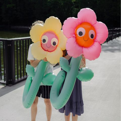 Due persone reggono grandi fiori gonfiabili con facce sorridenti, che nascondono i loro volti, mentre si trovano su un ponte sull'acqua.