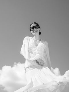Una donna in un ampio vestito bianco e grandi occhiali da sole sorride sotto un cielo nuvoloso; la foto è in bianco e nero.