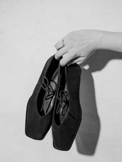 Una mano con un anello al dito tiene un paio di scarpe stringate nere con punta quadrata davanti a uno sfondo semplice e chiaro.