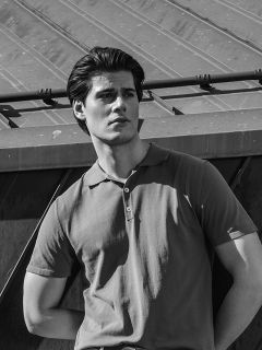 Un uomo in una polo a maniche corte è all'aperto, con lo sguardo rivolto di lato, sullo sfondo un tetto metallico inclinato. Foto in bianco e nero.