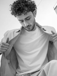Foto in bianco e nero di un uomo con capelli ricci che guarda in basso; indossa una giacca leggera sopra una semplice maglietta bianca e tiene la giacca aperta.