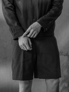 Foto in bianco e nero di una persona in camicia e pantaloncini, con le mani incrociate davanti alla vita, in piedi davanti a un muro strutturato.