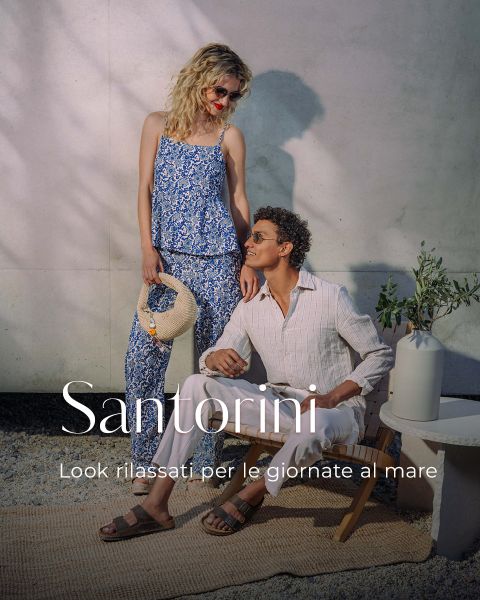 Una donna in un outfit blu a fiori e un uomo in abiti chiari posano davanti a una parete, sulla quale sono visualizzati "Santorini" e un testo italiano sull'aspetto rilassato al mare.