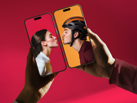 Zwei Hände halten Smartphones, auf deren Bildschirmen eine Frau und ein Mann mit Helm zu sehen sind, die sich vor bunten Hintergründen scheinbar durch die Bildschirme küssen.