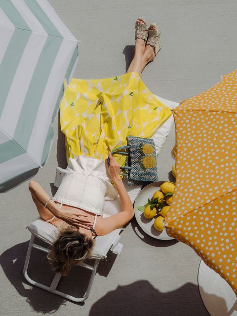 Una donna in gonna gialla siede su una sedia sotto ombrelloni, con limoni in una ciotola e una borsa lì vicino su una terrazza soleggiata.