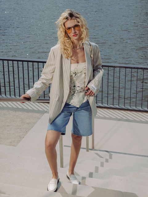 Una donna che indossa occhiali da sole, un blazer a righe, un top floreale, shorts di jeans e scarpe bianche, sta in piedi su un lungomare con una ringhiera dietro di lei.