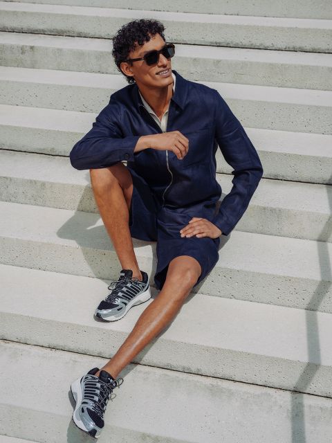Un uomo in pantaloncini e giacca blu navy, occhiali da sole e scarpe da ginnastica è seduto su una scala di cemento all'aperto, sorride e si rilassa sotto il sole splendente.