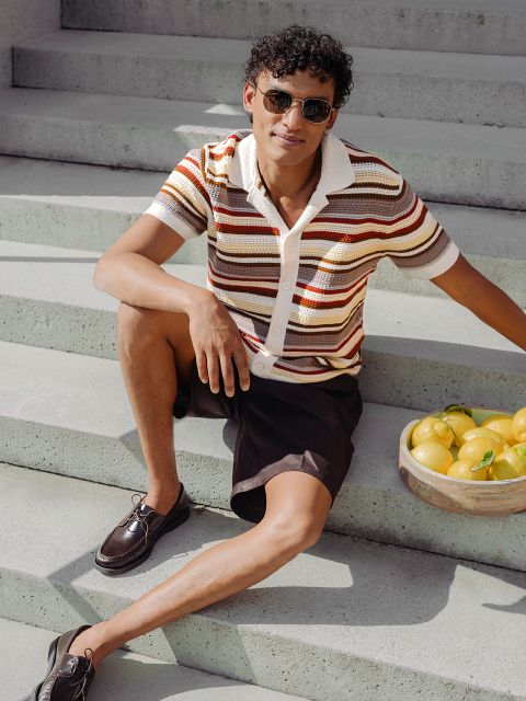 Un uomo con occhiali da sole e una camicia a righe siede su una scalinata esterna accanto a un cesto di limoni e sorride con disinvoltura alla luce del sole.