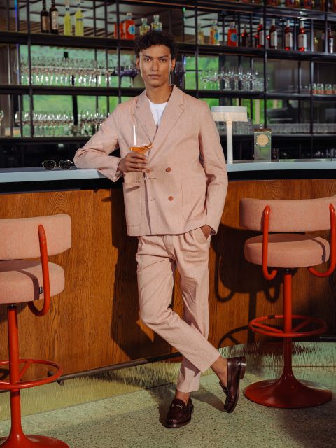 Un uomo in un abito rosa chiaro con una camicia bianca è in piedi a un bar con un drink in mano, con sgabelli da bar rosa e bicchieri sullo sfondo.