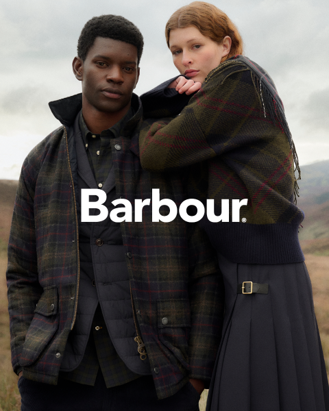 Un uomo e una donna con giacche Barbour a quadri posano all'aperto, vicini, davanti a un cielo nuvoloso e a un paesaggio collinare. Il logo Barbour è posizionato al centro.