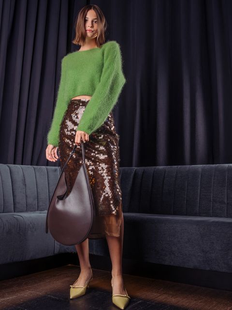 Una donna posa in un soffice maglione verde, una gonna midi marrone con paillettes e décolleté giallo chiaro, e tiene in mano una grande borsa hobo in pelle marrone scuro davanti a una tenda scura.