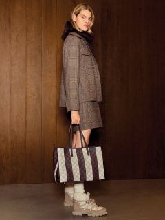 Una donna con un outfit a quadri tiene in mano una borsa a quadri, si trova in un interno davanti a una parete di legno e indossa stivali stringati.