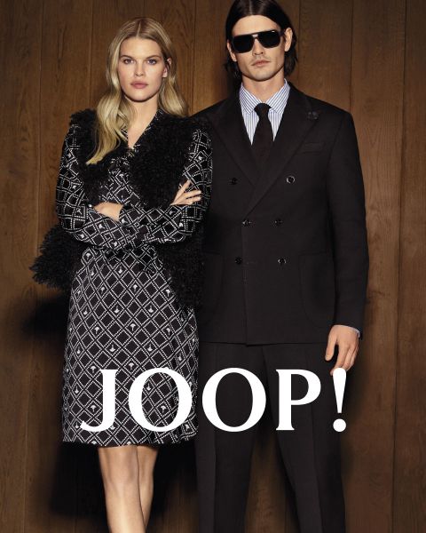 Una donna in un vestito a fantasia e un uomo in un abito nero stanno fianco a fianco davanti a una parete di legno, sulla quale è scritto "JOOP!" in lettere bianche e in grassetto.
