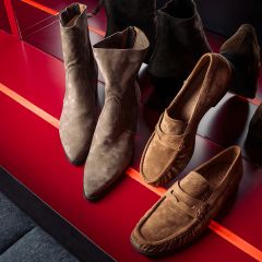 Due paia di scarpe di camoscio marroni - stivaletti e mocassini - esposte su uno scaffale rosso lucido, in cui si riflettono i loro riflessi.