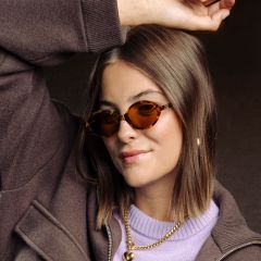 Una donna con capelli castani, che indossa occhiali da sole con montatura tartarugata, una giacca marrone, un top viola e collane d'oro, sorride e alza un braccio.