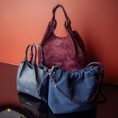 Tre eleganti borse - una bordeaux, una blu navy con catena e una grigio scuro - sono state presentate davanti a una parete rossastra su una superficie riflettente.