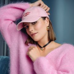 Una donna con un soffice maglione rosa e un cappello rosa posa in una stanza; indossa orecchini d'oro e un girocollo nero con un ciondolo a forma di cuore turchese.
