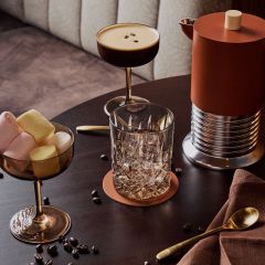Un tavolo con un cocktail, un bicchiere, una ciotola di marshmallow, una caffettiera, un cucchiaino d'oro, un tovagliolo e chicchi di caffè sparsi.