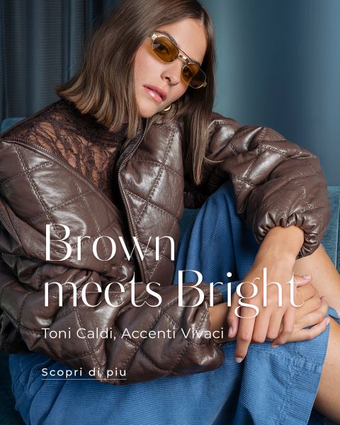Una donna con una giacca trapuntata marrone, un top di pizzo, una gonna blu e occhiali da sole gialli è seduta, mentre sullo sfondo è visibile un testo in lingua italiana che promuove la moda.