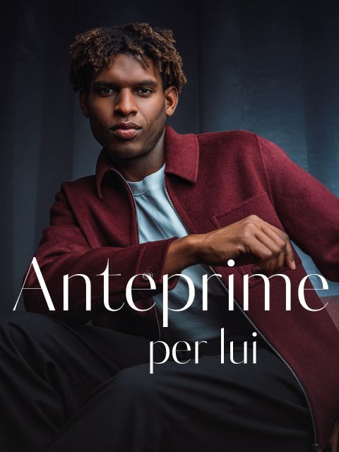 Un uomo con una giacca bordeaux posa con sicurezza davanti a uno sfondo scuro. Sull'immagine si estende la scritta bianca "Anteprime per lui".