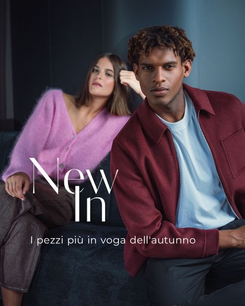 Due modelle con outfit autunnali eleganti sono sedute su un divano scuro. Il testo recita: „New In. I pezzi più in voga dell’autunno“.