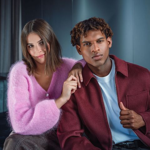 Zwei Menschen sitzen eng beieinander, einer trägt einen rosa Pullover, der andere eine weinrote Jacke; beide blicken mit entspannter Miene in die Kamera.