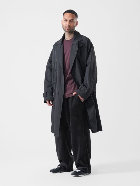 Ein Mann, der einen schwarzen Trenchcoat, ein weinrotes Hemd, eine schwarze Hose mit weitem Bein und schwarze Sneaker trägt, steht vor einem schlichten weißen Hintergrund.