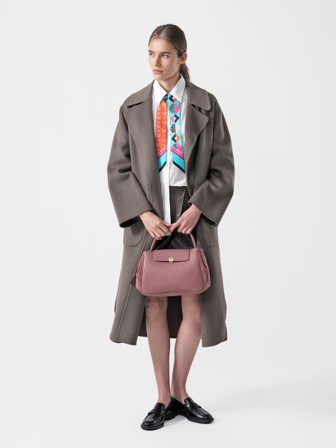 Eine Frau in einem langen braunen Mantel, einem bunten Schal, einer weißen Bluse, einem braunen Rock und schwarzen Loafern, die eine rosa Handtasche in der Hand hält und vor einem schlichten weißen Hintergrund steht.